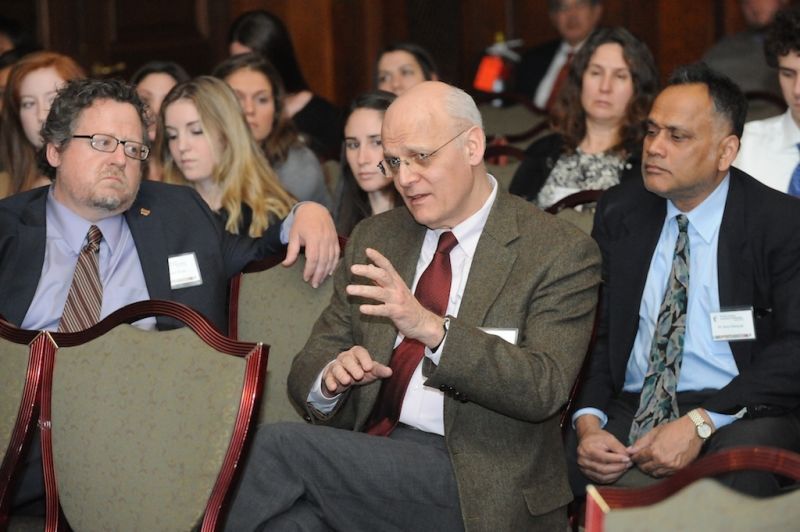 A group of people are seated in a conference room. Three men in the front row are engaged in discussion, with the man in the center gesturing as he speaks. Others in the background appear to be listening or observing. The setting suggests a formal event or seminar.